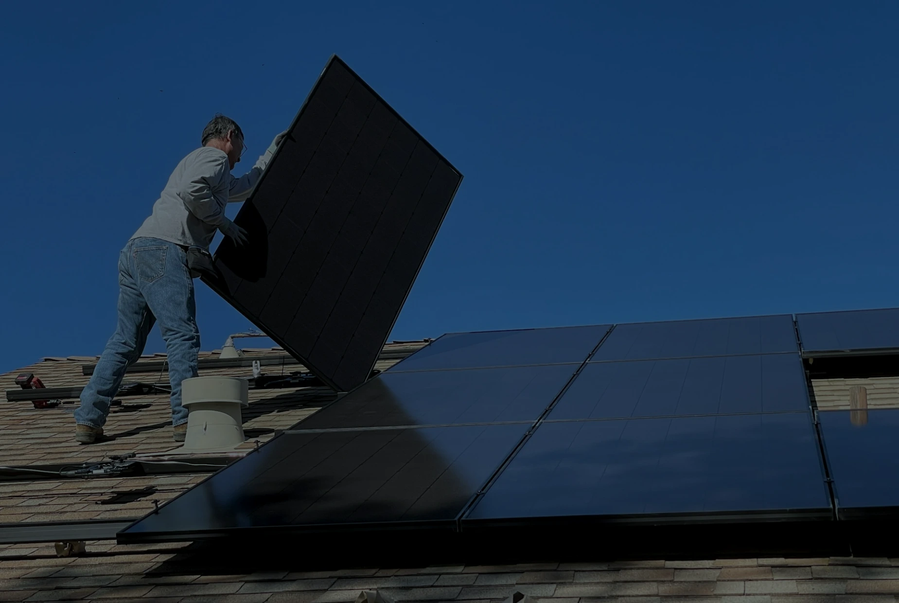 A person installing a black solar panel on a shingled roof under blue sky